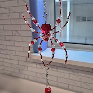 May include: A decorative spider ornament made of red and clear beads. The spider has a heart-shaped body and is suspended by a silver chain. The legs are made of red beads and clear crystal beads. The ornament is hanging in front of a window.