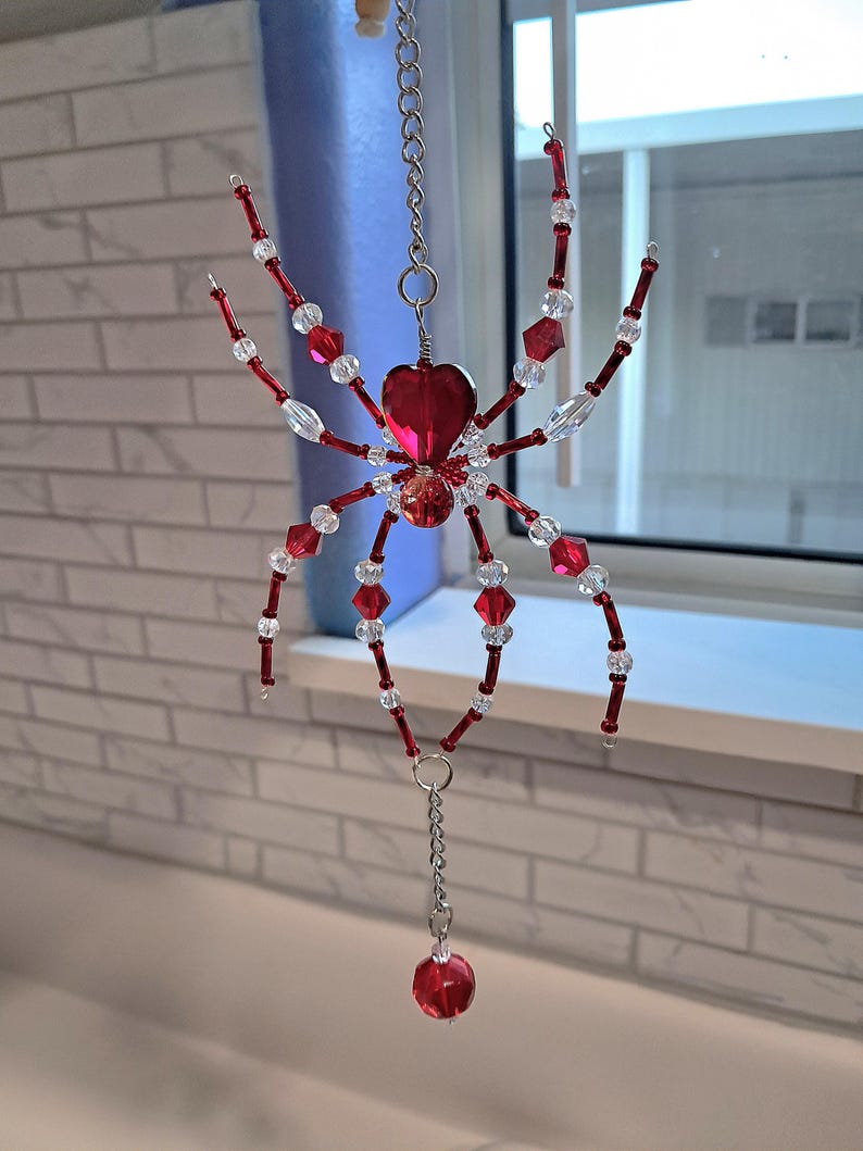 May include: A decorative spider ornament made of red and clear beads. The spider has a heart-shaped body and is suspended by a silver chain. The legs are made of red beads and clear crystal beads. The ornament is hanging in front of a window.