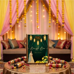 May include: A framed sign with the words "Bangle Bar" in gold script, featuring a woman in traditional attire. The sign is on a gold table with colorful bangles. The background includes draped fabric and decorative lights.