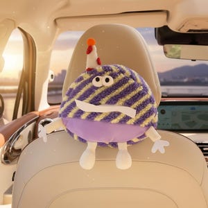 May include: A plush, whimsical car headrest cover shaped like a cartoon character. The character has a purple and yellow striped body, white eyes, a white mustache, and a party hat. It has white arms and feet. The headrest cover is designed to fit over a car seat headrest.