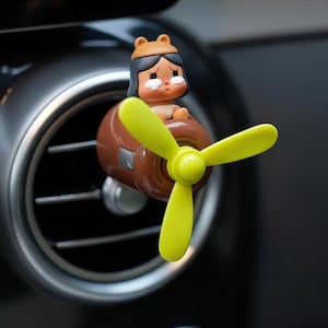 Könnte beinhalten: Ein brauner und gelber Auto-Lufterfrischer in Flugzeugform mit einem Cartoon-Charakter als Pilot. Der Propeller ist gelb und der Pilot trägt einen braunen Hut. Der Lufterfrischer ist an einer Lüftungsöffnung im Auto befestigt.