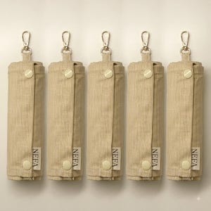 Op de afbeelding: Vijf cilindervormige stoffen etuis met een beige en wit krijtstreepmotief. Elk etui is voorzien van een goudkleurige metalen karabijnhaak aan de bovenkant, twee crèmekleurige drukknopen en een klein wit stoffen label met de tekst NEFA.
