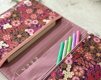 May include: Open book cover with a floral pattern in shades of purple, pink, and white. The cover is open, revealing a book and several colorful pens. A zipper closure secures the cover. The book cover is a stylish accessory for book lovers.