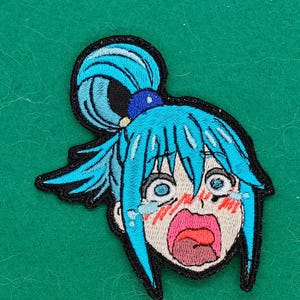 May include: An embroidered patch featuring the head of an anime character with bright blue hair styled in a high ponytail. The character has a wide-open mouth, large blue eyes with tears, and red blushing cheeks, all outlined in black thread.