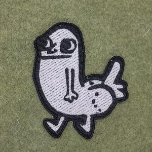 May include: A white and black embroidered patch featuring a cartoonish creature with a large head, small arms, and a rounded body. The patch has a black outline and is set against a green background.