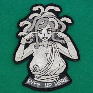 May include: Embroidered patch featuring a stylized Medusa figure with snakes for hair, pointing at her eyes. The patch is in shades of gray and white, with the text "EYES UP HERE" at the bottom. The patch has a black border.