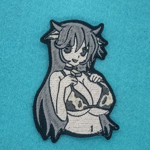 May include: Embroidered patch featuring a cartoon cow-girl character with long hair, horns, and a cow-print bikini top. The patch is primarily black, white, and gray, with a black border. The background is a light blue fabric.
