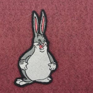 May include: Embroidered patch featuring a cartoon rabbit character with gray fur, pink inner ears, and a white belly. The patch has a black border and is set against a textured maroon background.