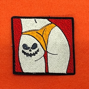 May include: Embroidered patch featuring a white backside wearing a yellow thong, with a jack-o'-lantern face embroidered on the buttocks. The patch is framed in a red border with a black outline, set against an orange background.