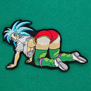 May include: Embroidered patch featuring a cartoon character in a kneeling pose. The character has blue hair, a white shirt, red shorts, and green leggings. The patch is outlined in black and has a green background.