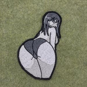 May include: Embroidered patch featuring a stylized anime-style figure in a suggestive pose. The patch has a black outline with gray and white fill, depicting the figure's form. The patch is on a green fabric background.