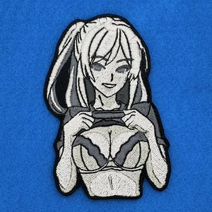 May include: Embroidered patch featuring a grayscale anime-style character with long hair in a ponytail. The character is depicted holding up their top, revealing a detailed bra. The patch has a black border and is set against a blue background.