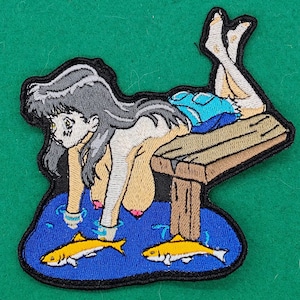 May include: Embroidered patch featuring an anime-style figure lounging on a wooden dock above blue water with two yellow fish. The figure has gray hair, blue shorts, and is in a relaxed pose. The patch has a black border.