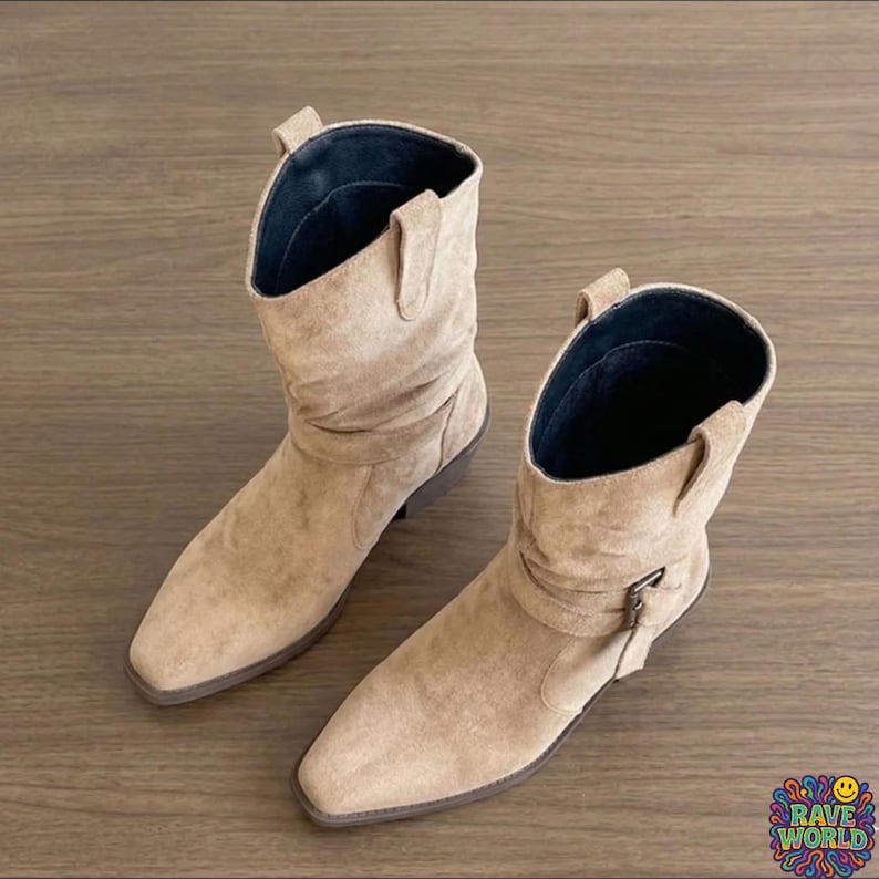 A close-up shot of stylish tan sheepskin and microfiber slouch boots worn at an outdoor music festival. The boots feature a soft, ruched shaft, a pointed toe, and a decorative wrap-around buckle strap. Cream-colored ribbed socks peek out from the top. The background shows a dusty festival ground with a blurred Ferris wheel and crowds under golden hour sunlight, emphasizing a bohemian, desert-chic aesthetic and durable, comfortable design.