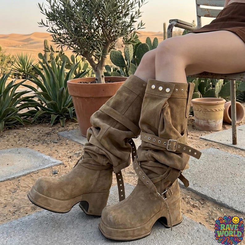 A close-up of a person walking in a desert landscape wearing tall, slouchy, tan microfiber leather boots. The boots feature a rugged design with multiple straps, brass buckles, and stud detailing on a thick platform sole. The person is styled in light-wash denim jeans and a leather wristband. The background shows a sunlit trail with Joshua trees, cacti, and distant mesas, highlighting the boots' durable, adventurous, and boho-western aesthetic.