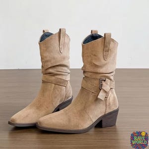 A close-up shot of stylish tan sheepskin and microfiber slouch boots worn at an outdoor music festival. The boots feature a soft, ruched shaft, a pointed toe, and a decorative wrap-around buckle strap. Cream-colored ribbed socks peek out from the top. The background shows a dusty festival ground with a blurred Ferris wheel and crowds under golden hour sunlight, emphasizing a bohemian, desert-chic aesthetic and durable, comfortable design.