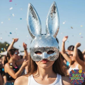 May include: A silver disco ball bunny mask with long ears. The mask covers the eyes and nose, with a black strap. The image includes the text "RAVE WORLD" in a colorful, psychedelic font, and people in the background.