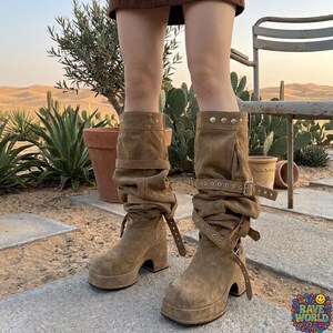 A close-up of a person walking in a desert landscape wearing tall, slouchy, tan microfiber leather boots. The boots feature a rugged design with multiple straps, brass buckles, and stud detailing on a thick platform sole. The person is styled in light-wash denim jeans and a leather wristband. The background shows a sunlit trail with Joshua trees, cacti, and distant mesas, highlighting the boots' durable, adventurous, and boho-western aesthetic.