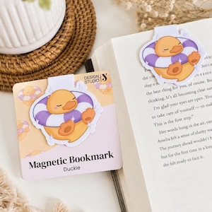 Op de afbeelding: Magnetische boekenlegger met een cartoon eend in een paarse en witte zwemband. De boekenlegger is gelabeld "Duckie" en staat op een licht perzikkleurige achtergrond. Design Studio Y is afgebeeld.