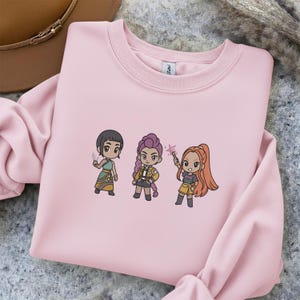 May include: A light pink sweatshirt featuring three cartoon characters. The characters are in a chibi style, each holding a different item. The sweatshirt is made of a soft material and has a crew neck.