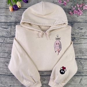 May include: Cream-colored hoodie with a drawstring hood and long sleeves. The front features a cartoon illustration of a gray creature with a smaller face and a black creature with a heart on the sleeve.