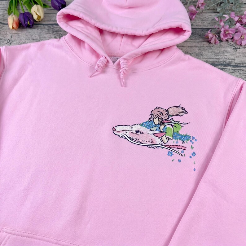 May include: A light pink hoodie with a drawstring hood and a kangaroo pocket. The hoodie features an embroidered design of a girl riding a white dragon with blue flowers.