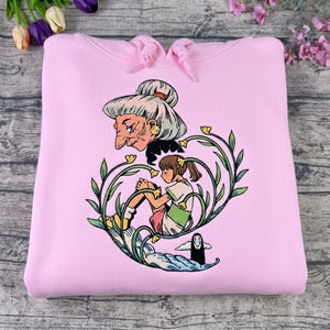 Puede incluir: Sudadera con capucha rosa claro con un dise&ntilde;o gr&aacute;fico colorido de la pel&iacute;cula de animaci&oacute;n El Viaje de Chihiro. El dise&ntilde;o incluye a Yubaba, Chihiro y Sin Cara, rodeados de elementos florales. La sudadera tiene una capucha con cord&oacute;n.