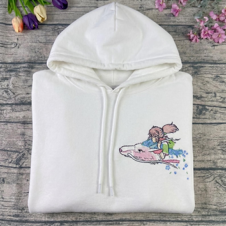 May include: White hooded sweatshirt with a drawstring and a colorful embroidered design. The design features a character riding a pink dragon with blue floral accents. The hoodie is made of a soft material.