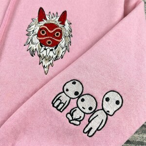 May include: Pink hoodie with embroidered designs. Features a red and white mask design on the chest and three white Kodama spirits on the sleeve. The hoodie is made of a soft, comfortable material.