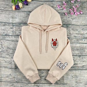 May include: A beige hoodie with a drawstring hood and long sleeves. The hoodie features an embroidered red and white mask design on the chest and three small white figures on the left sleeve.