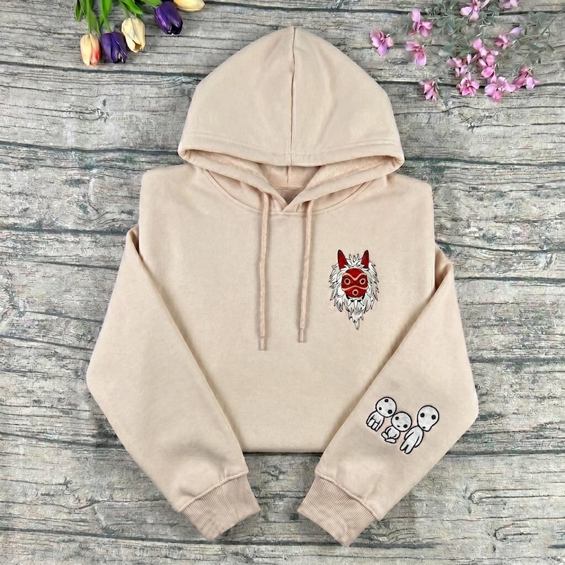 May include: A beige hoodie with a drawstring hood and long sleeves. The hoodie features an embroidered red and white mask design on the chest and three small white figures on the left sleeve.