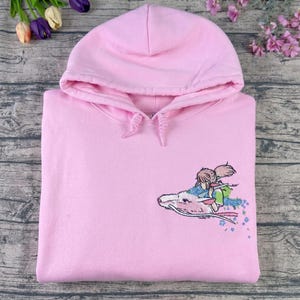 May include: A light pink hoodie with a drawstring hood. The hoodie features an embroidered design of a character riding a white dragon. The design includes shades of blue, green, and pink. The hoodie is made of a soft material.