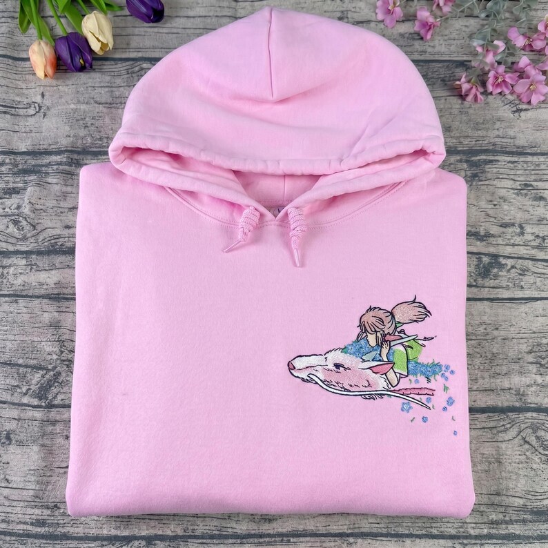 May include: A light pink hoodie with a drawstring hood. The hoodie features an embroidered design of a character riding a white dragon. The design includes shades of blue, green, and pink. The hoodie is made of a soft material.