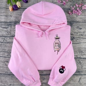 May include: A light pink hoodie with a drawstring hood and long sleeves. The front features a cartoon illustration of a gray mouse-like creature with a smaller face. The left sleeve has a black, fluffy creature with a red heart.