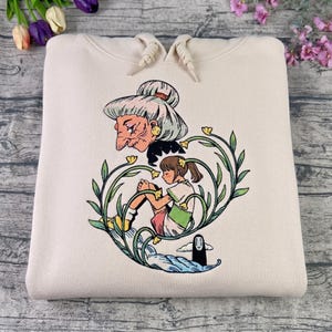 Puede incluir: Sudadera con capucha de color crema con una colorida ilustraci&oacute;n de una pel&iacute;cula de animaci&oacute;n. El dise&ntilde;o incluye un personaje con pelo gris, una ni&ntilde;a y una criatura, todo rodeado de enredaderas verdes y flores amarillas. La sudadera tiene una capucha con cord&oacute;n.