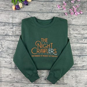 May include: A dark green sweatshirt with the text "THE NIGHT CRAWLERS" in orange and white script. Below the text is the phrase "THE WEIRDEST & WILDEST OF THEM ALL". The sweatshirt is made of a soft material.