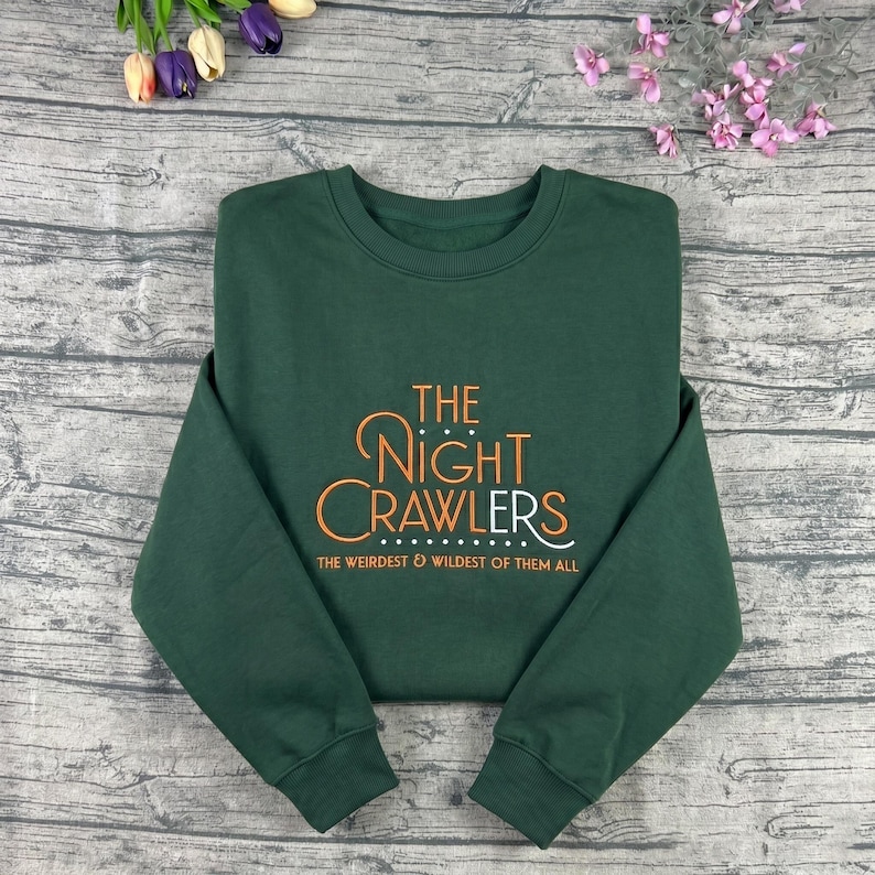 May include: A dark green sweatshirt with the text "THE NIGHT CRAWLERS" in orange and white script. Below the text is the phrase "THE WEIRDEST & WILDEST OF THEM ALL". The sweatshirt is made of a soft material.