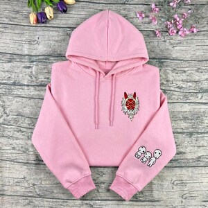 May include: A light pink hoodie with a drawstring hood. The hoodie features an embroidered design of a red and white mask on the chest and three white spirit figures on the left sleeve. The hoodie is made of a soft material.