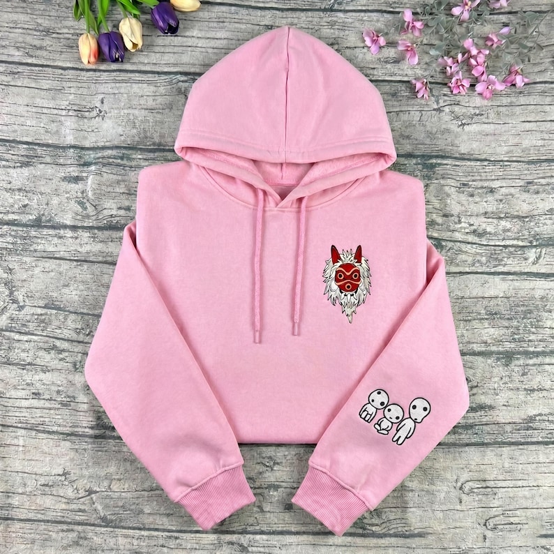 May include: A light pink hoodie with a drawstring hood. The hoodie features an embroidered design of a red and white mask on the chest and three white spirit figures on the left sleeve. The hoodie is made of a soft material.