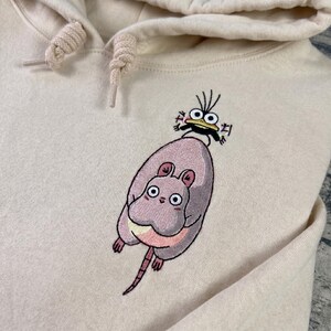 May include: Cream-colored hoodie with an embroidered design featuring a large, pink, cartoon-style rat with a small frog on its back. The rat has large eyes and a long tail. The frog has large eyes and a black mustache.