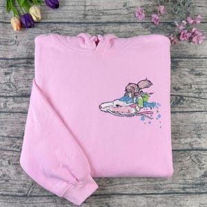 May include: A light pink hoodie with an embroidered design. The design features a character riding a white dragon with blue and pink floral accents. The hoodie has a hood and long sleeves.