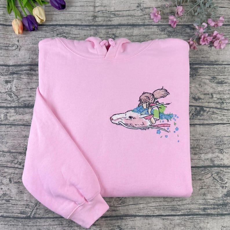 May include: A light pink hoodie with an embroidered design. The design features a character riding a white dragon with blue and pink floral accents. The hoodie has a hood and long sleeves.