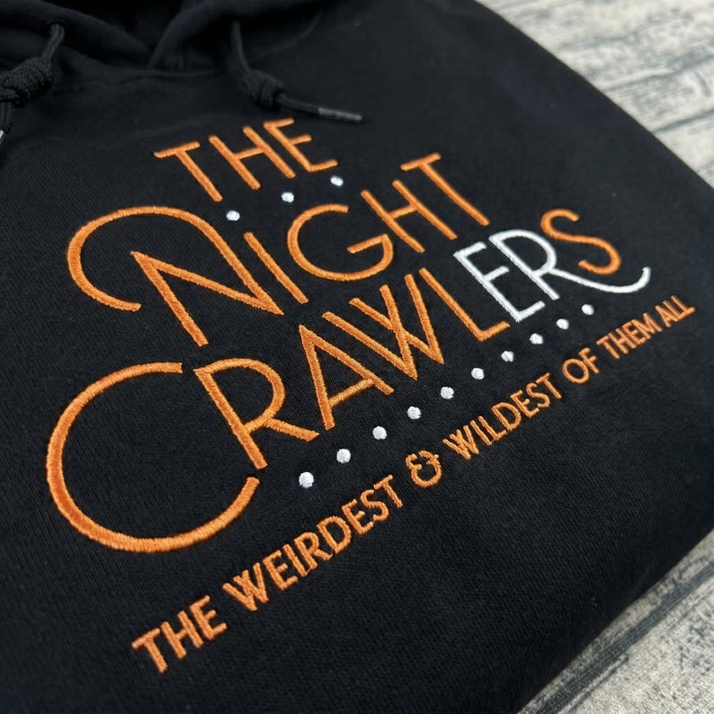 May include: Black hooded sweatshirt with the embroidered text "THE NIGHT CRAWLERS" in orange and white. The phrase "THE WEIRDEST & WILDEST OF THEM ALL" is embroidered below the main text. The sweatshirt has a drawstring hood.