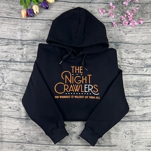 May include: Black hooded sweatshirt with the text "THE NIGHT CRAWLERS" in orange and white script. The phrase "THE WEIRDEST & WILDEST OF THEM ALL" is printed below the main text. The sweatshirt is displayed on a wooden surface.
