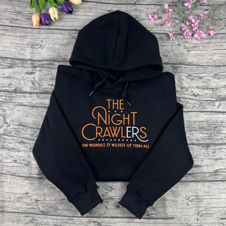 May include: Black hooded sweatshirt with the text "THE NIGHT CRAWLERS" in orange and white script. The phrase "THE WEIRDEST & WILDEST OF THEM ALL" is printed below the main text. The sweatshirt is displayed on a wooden surface.