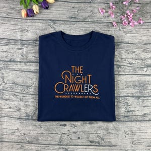 May include: Navy blue t-shirt with the text "THE Night Crawlers" in orange script. Below the text is the phrase "THE WEIRDEST & WILDEST OF THEM ALL" in white. The shirt is folded on a light gray surface.