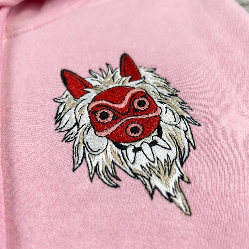 May include: Pink hoodie featuring an embroidered design of a red and white wolf-like mask. The mask has a detailed design with red ears and a red face mask with black eye details. The embroidery is on the upper left chest area of the hoodie.