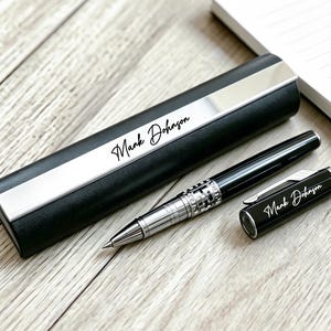 May include: A black and silver pen set with the name "Mark Johnson" in cursive script. The set includes a pen, pen case, and pen cap, all resting on a wooden surface next to a spiral notebook.