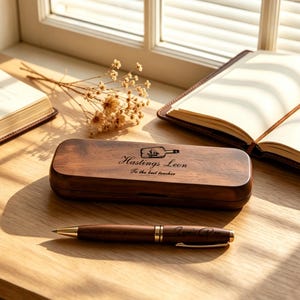 May include: A dark brown wooden pen case with a hinged lid featuring a laser-engraved ship in a bottle icon, the name Hastings Leon, and the text To the best teacher. A matching dark brown wooden ballpoint pen with gold accents lies in front.