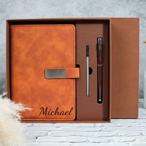May include: A brown leather journal with a metal clasp and the name "Michael" engraved on the cover. The journal is packaged in a brown box with a matching pen and refill.